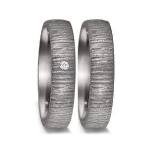 Nahaufnahme zweier ineinander verschlungener Eheringe aus einem dunklen, grau wirkenden Metall vor einem neutralen Hintergrund. Die Oberfläche der Ringe ist von einer markanten, rustikalen und leicht unebenen Textur geprägt, die an Rindenstruktur oder eine gehammerte Oberfläche erinnert. Der linke Ring ist mit einem einzelnen, mittig eingesetzten Brillanten akzentuiert, während der rechte Ring schlicht gehalten ist.