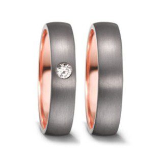 Nahaufnahme eines Paares moderner Eheringe vor neutralem Hintergrund. Die Ringe haben ein zweifarbiges Design mit einer dunkelgrauen, matt gebürsteten Außenseite und einer glänzend polierten Innenseite aus Roségold. Der linke Ring ist mit einem einzelnen, mittig gefassten Brillanten akzentuiert, während der rechte Ring schlicht gehalten ist.