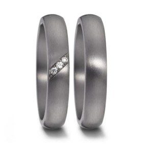 Nahaufnahme eines Paares moderner Eheringe aus dunkelgrauem, mattiertem Metall vor einem neutralen Hintergrund. Der linke Ring ist mit drei kleinen, diagonal angeordneten Brillanten verziert, die in einer feinen Linie eingelassen sind. Der rechte Ring ist schlicht und schmal gehalten, passend zum Design des linken Rings.
