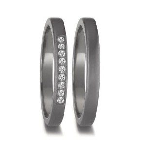 Nahaufnahme zweier ineinander verschlungener Eheringe aus einem dunklen, grau wirkenden Metall mit mattierter Oberfläche vor einem neutralen Hintergrund. Der linke Ring ist mit einer vertikalen Reihe von acht kleinen, funkelnden Brillanten verziert, die in einer eingelassenen Schiene angeordnet sind. Der rechte Ring ist schlicht und minimalistisch gehalten.