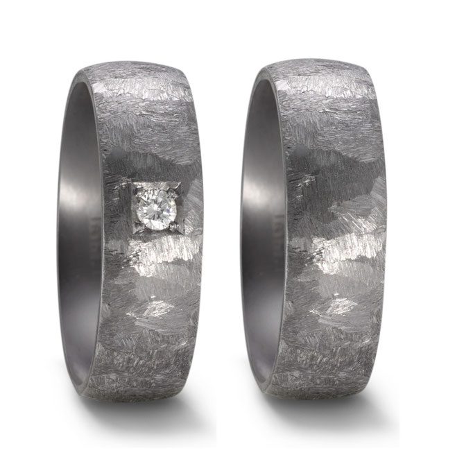 Ein archaischer Ausdruck von Verbundenheit: Diese Eheringe bestechen durch ihre markante, tief strukturierte Oberfläche in dunklem Metall, die einen spannenden Kontrast zum eleganten Funkeln des einzelnen Diamanten bildet. Ein Design für Paare, die eine moderne, individuelle und rustikale Interpretation klassischer Trauringe suchen.