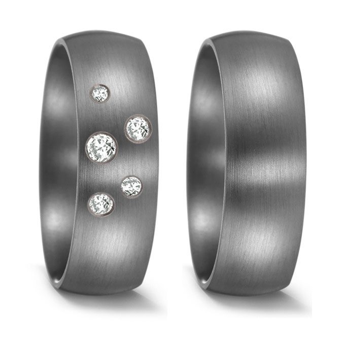 Ein Hauch von Sternenhimmel: Dieses Trauring-Set besticht durch die kühle, matte Eleganz des dunklen Metalls, das als perfekter Hintergrund für die verspielte, asymmetrische Anordnung der Diamanten auf dem einen Ring dient. Ein Design für Paare, die eine moderne und poetische Interpretation klassischer Eheringe suchen.