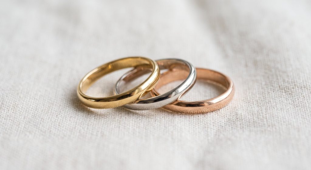 Eine Nahaufnahme von drei schlichten, polierten Metallringen, die ineinandergreifen und auf einem hellbeigen, grob gewebten Leinenstoff liegen. Von links nach rechts sind die Ringe in Gelbgold, Weißgold (oder Platin) und Roségold gehalten. Die Ringe sind im Tri-Color-Stil arrangiert und werfen weiche Schatten auf die Stofftextur.