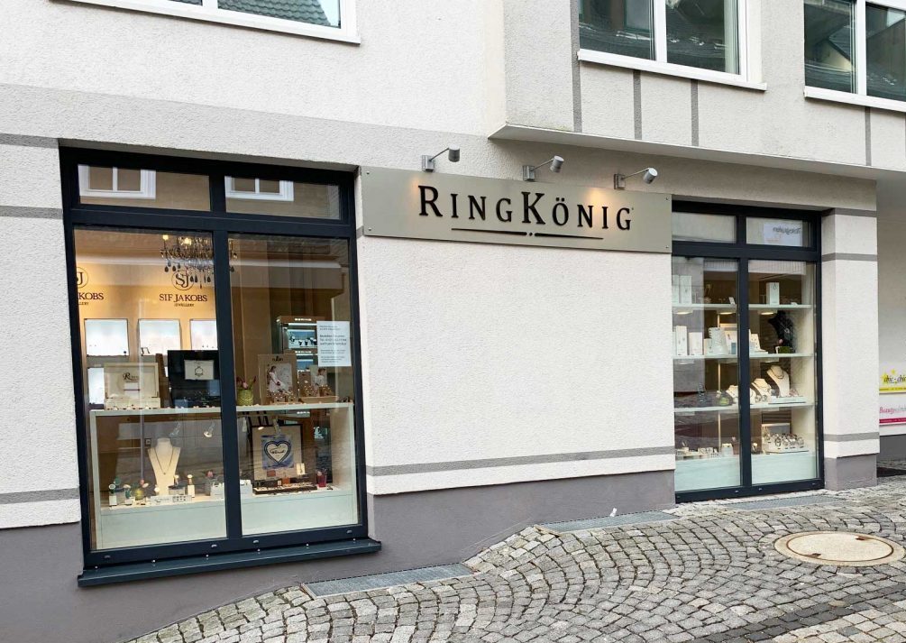 Tageslichtaufnahme der Fassade des Juweliergeschäfts „RingKönig“. Über dem Eingang ist der Schriftzug „RINGKÖNIG“ auf einem Schild montiert. Große, bodentiefe Schaufenster präsentieren verschiedene Schmuckstücke auf eleganten Displays. Vor dem Geschäft erstreckt sich ein gepflasterter Gehweg.