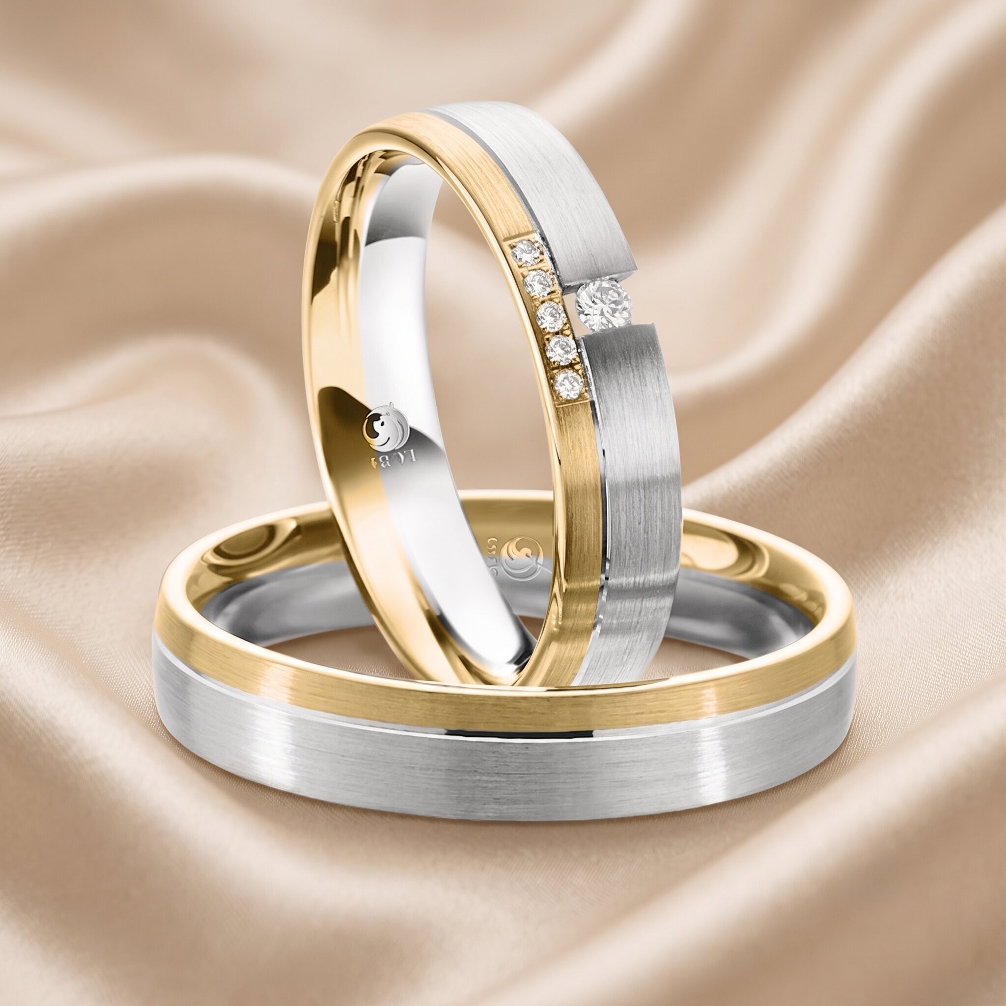 Ein Paar Bicolor-Trauringe aus gebürstetem Weißgold und poliertem Gelbgold, elegant arrangiert auf weichem, drapiertem champagnerfarbenem Satinstoff. Der schräg stehende Damenring ist mit einer Reihe kleiner Diamanten im Brillantschliff verziert. Ein graviertes Logo ist auf der Innenseite des Damenrings sichtbar.