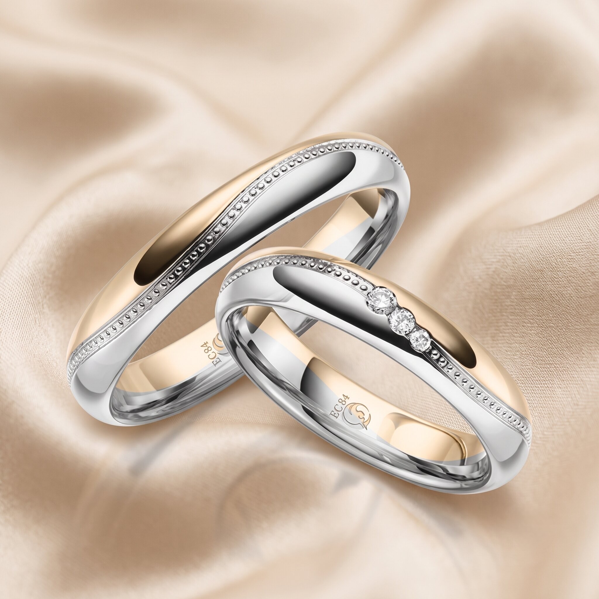 Eine Makroaufnahme von zwei ineinander verschlungenen Bicolor-Eheringen aus Silber und Gold, die auf einem weich drapierten champagnerfarbenen Satinhintergrund liegen. Der linke, breitere Ring ist schlicht und bicolor mit Perlenrandverzierungen. Der rechte Ring hat ebenfalls einen Perlenrand, ist aber mit drei Kanalfassungsdiamanten besetzt. Auf den Innenseiten beider Ringe ist die Gravur 'EC84' und ein Symbol sichtbar.
