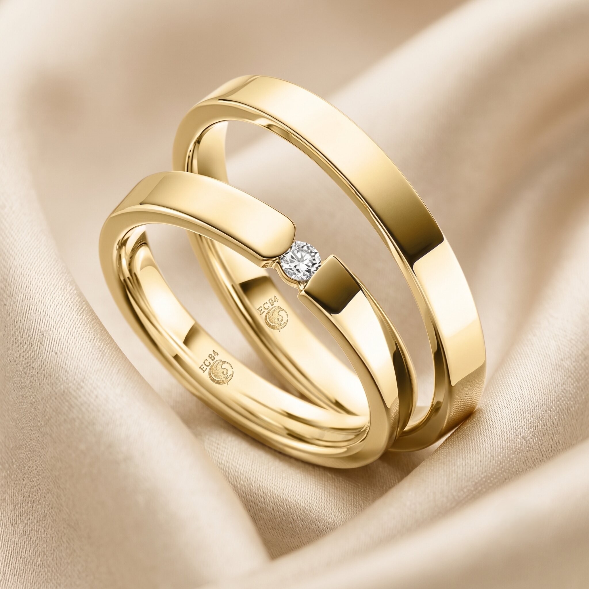 Zwei ineinander verschlungene, hochglanzpolierte goldene Eheringe mit flacher Schiene liegen auf einem luxuriösen, weich gefalteten champagnerfarbenen Satinstoff. Der vordere, schmalere Ring zeigt einen einzigen runden Brillanten (Diamanten), der bündig in der Mitte seiner Schiene gefasst ist. Auf der Innenseite beider Ringe ist deutlich eine Gravur mit dem "EC84"-Schwanenlogo und dem Schriftzug "EC84" zu sehen. Die Beleuchtung ist weich und diffus, und die Schärfentiefe ist gering, wodurch der Hintergrund weichgezeichnet ist und die Ringe im Fokus stehen.