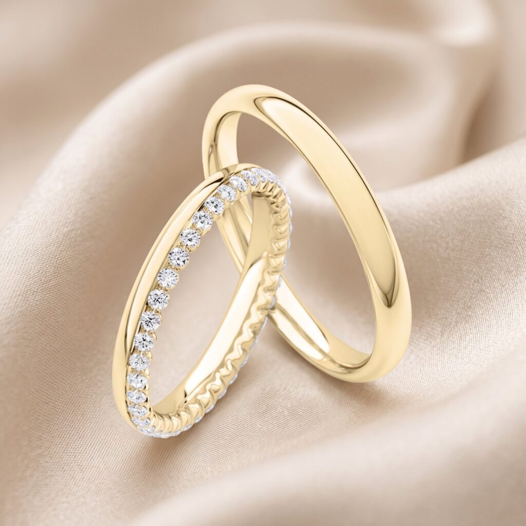Zwei ineinander verschlungene Eheringe aus Gelbgold auf drapiertem champagnerfarbenem Satin. Der linke Ring ist ein funkelnder Ewigkeitsring (Eternity-Ring), der vollständig mit kleinen runden Diamanten besetzt ist. Der rechte Ring ist ein klassischer, schlichter, hochglanzpolierter Goldreif. Nahaufnahme.