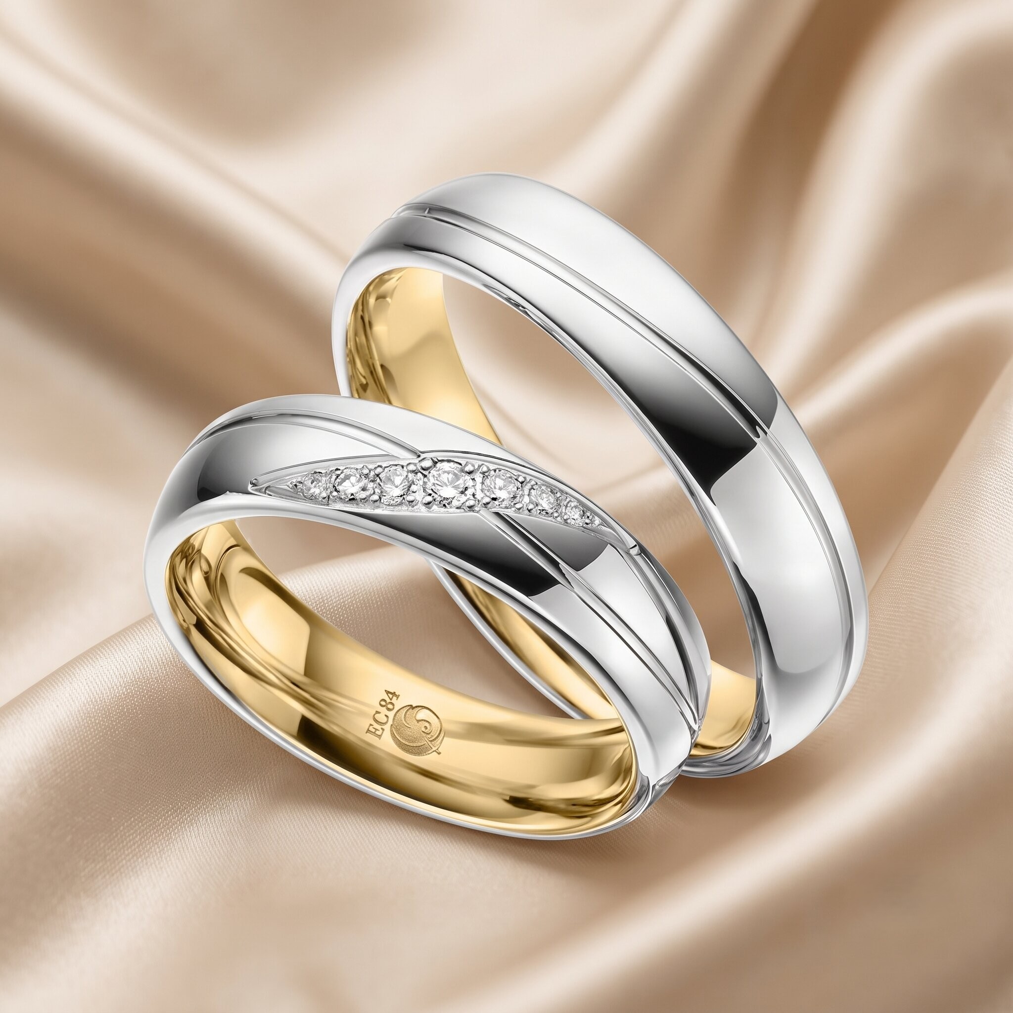 Nahaufnahme von zwei ineinander verschlungenen Bicolor-Trauringen aus poliertem Weißgold und Gelbgold, die auf einem weich fließenden, goldfarbenen Satinhintergrund ruhen. Der obere Ring ist ein schlichter Bändering aus Weißgold mit einer zentralen Nut. Der untere Ring hat ein diagonal verlaufendes Band, das mit mehreren kleinen, runden pavé-gefassten Diamanten besetzt ist, flankiert von goldenen Akzenten. Die Innenseite des unteren Rings ist goldfarben und zeigt deutlich die Gravur "EC 84" sowie ein kleines Markensymbol.