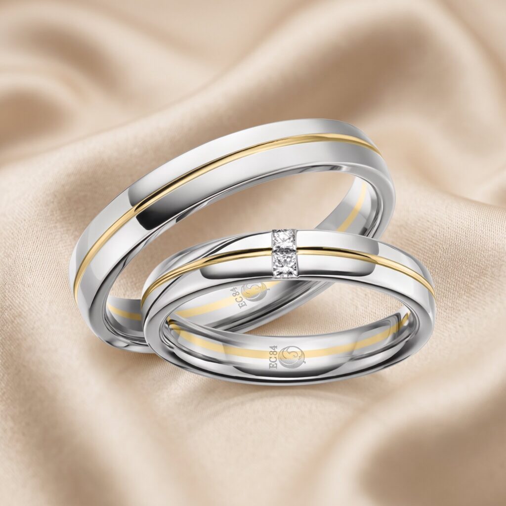 Nahaufnahme von zwei verschränkten Bicolor-Trauringen aus silber- und gelbgoldfarbenem Metall. Der vordere Ring ist mit drei Diamanten besetzt. Im Inneren sind Gravuren mit dem Text 'EC84' und einem Symbol sichtbar. Die Ringe ruhen auf drapiertem, champagnerfarbenem Stoff.