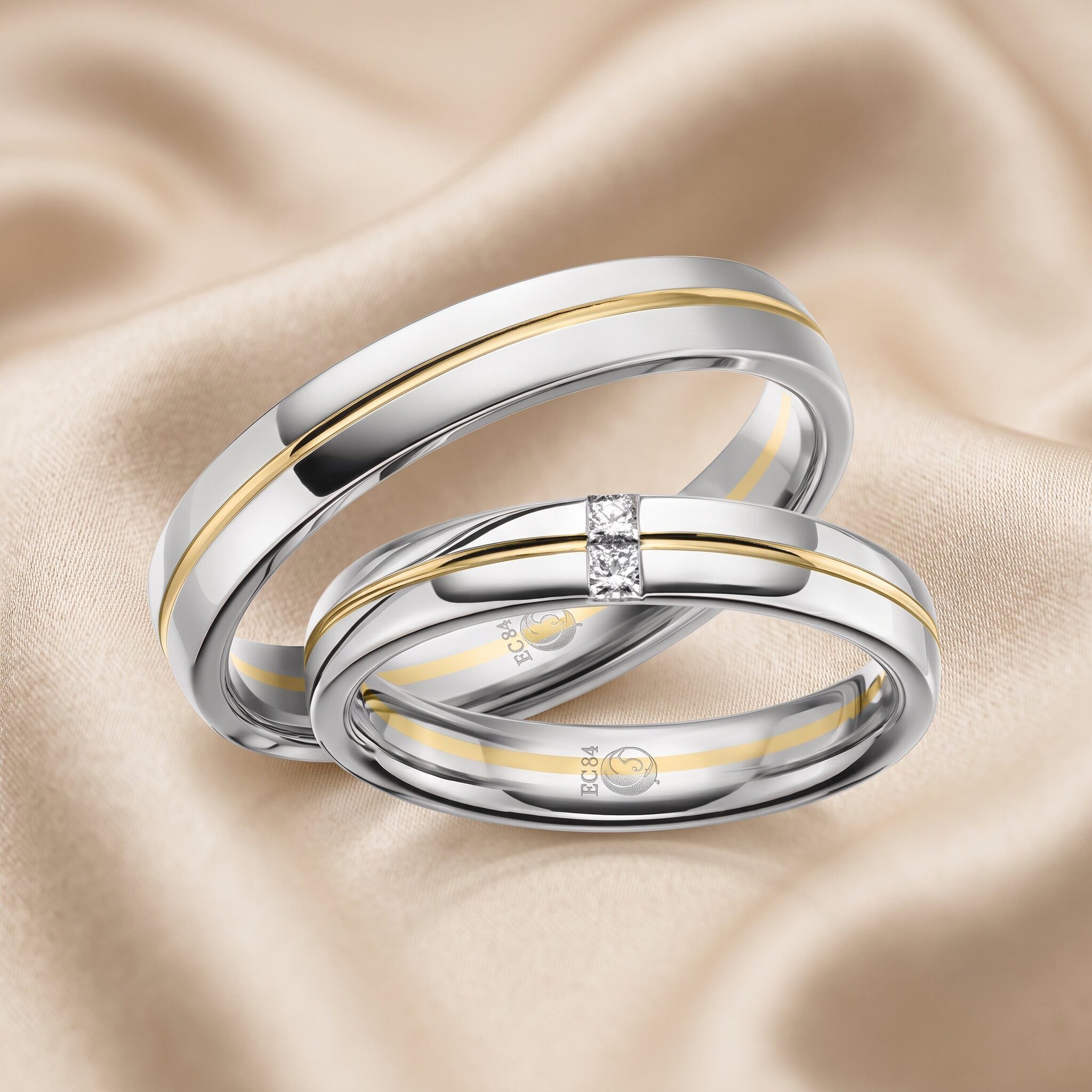 Nahaufnahme von zwei verschränkten Bicolor-Trauringen aus silber- und gelbgoldfarbenem Metall. Der vordere Ring ist mit drei Diamanten besetzt. Im Inneren sind Gravuren mit dem Text 'EC84' und einem Symbol sichtbar. Die Ringe ruhen auf drapiertem, champagnerfarbenem Stoff.