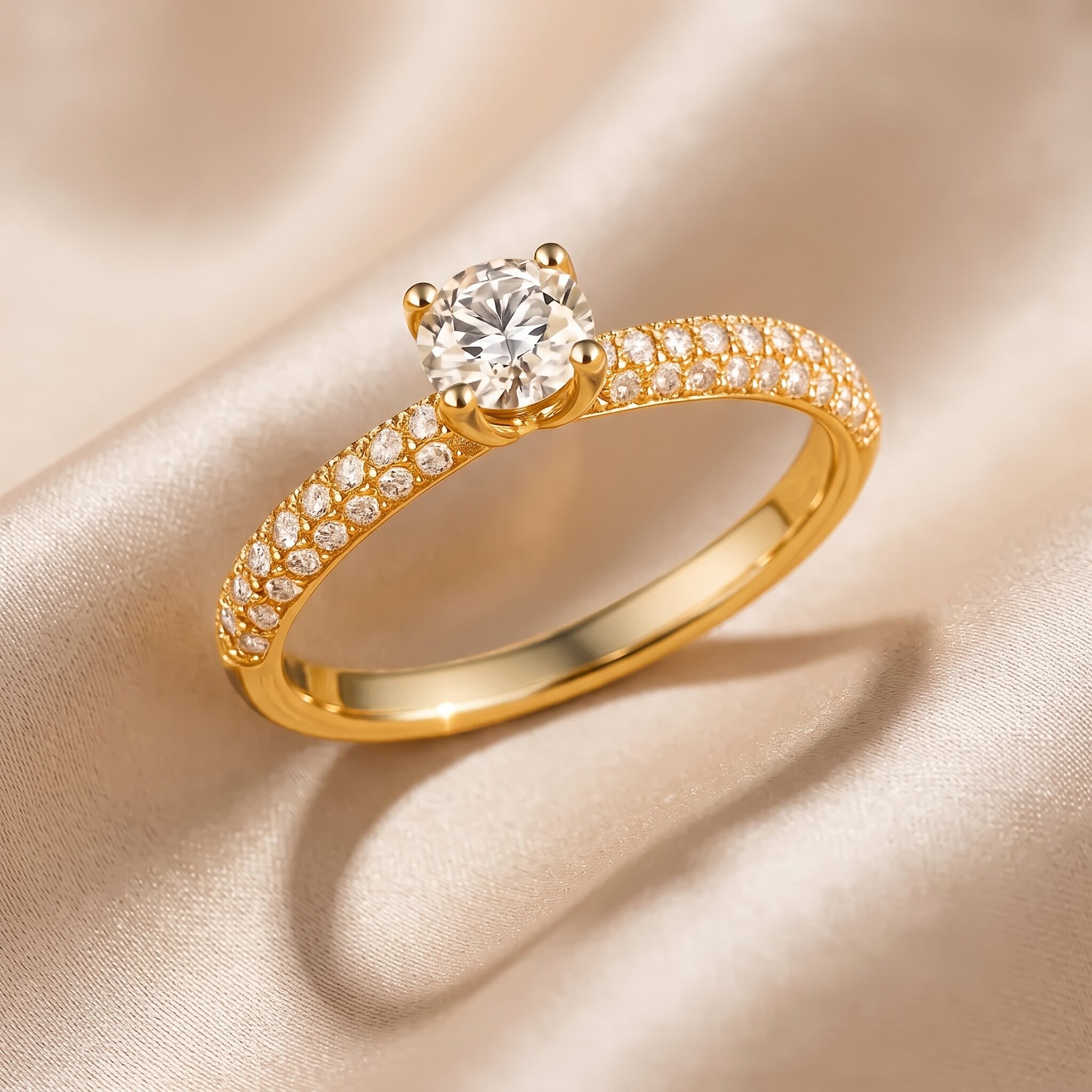 Ein eleganter Verlobungsring aus Gelbgold mit einem zentralen, runden Brillanten und einem mit kleinen Diamanten besetzten Ringband (Pavé-Fassung), der auf einem weichen, champagnerfarbenen Seidensatin-Hintergrund ruht.