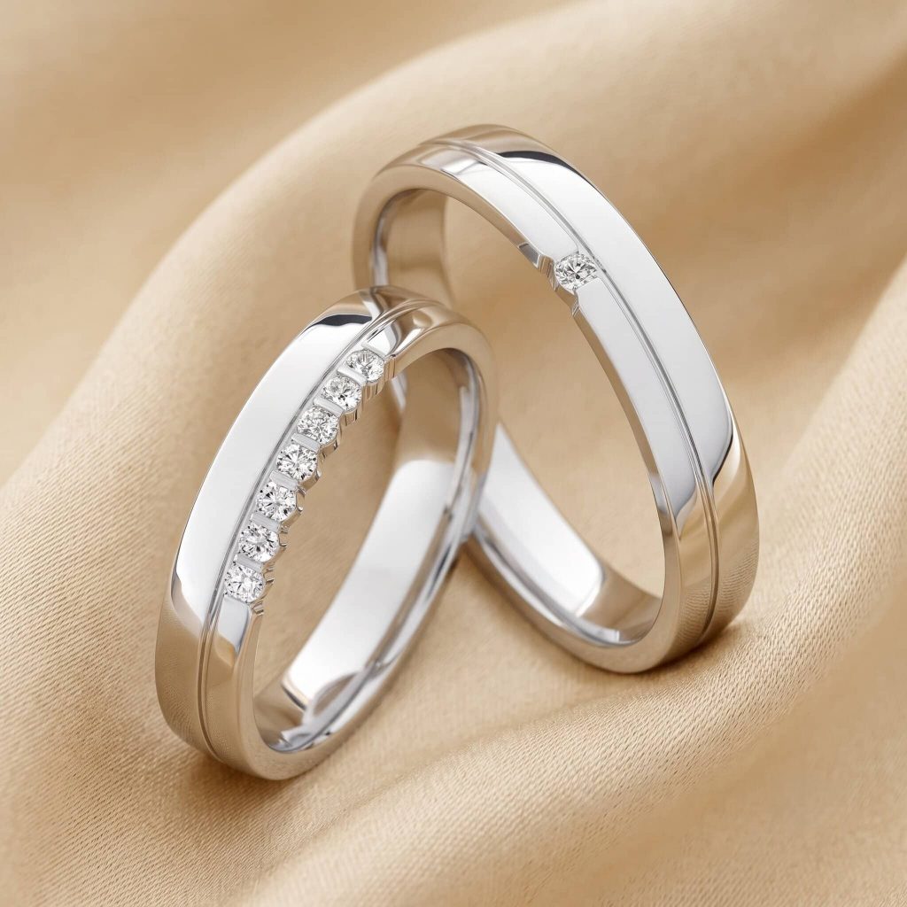 Nahaufnahme zweier ineinander verschlungener Eheringe aus kühlem Weißgold, die auf einem weich drapierten, champagnerfarbenen Stoff liegen. Der linke Ring ist mit einer Reihe kleiner Diamanten verziert, die in einer eingelassenen Schiene gefasst sind. Der rechte Ring hat ein ähnliches Design mit einer durchgehenden Rille, ist aber nur mit einem einzelnen kleinen Diamanten akzentuiert.