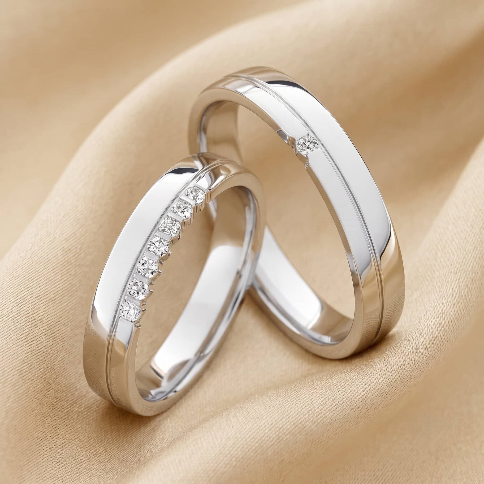 Nahaufnahme zweier ineinander verschlungener Eheringe aus kühlem Weißgold, die auf einem weich drapierten, champagnerfarbenen Stoff liegen. Der linke Ring ist mit einer Reihe kleiner Diamanten verziert, die in einer eingelassenen Schiene gefasst sind. Der rechte Ring hat ein ähnliches Design mit einer durchgehenden Rille, ist aber nur mit einem einzelnen kleinen Diamanten akzentuiert.