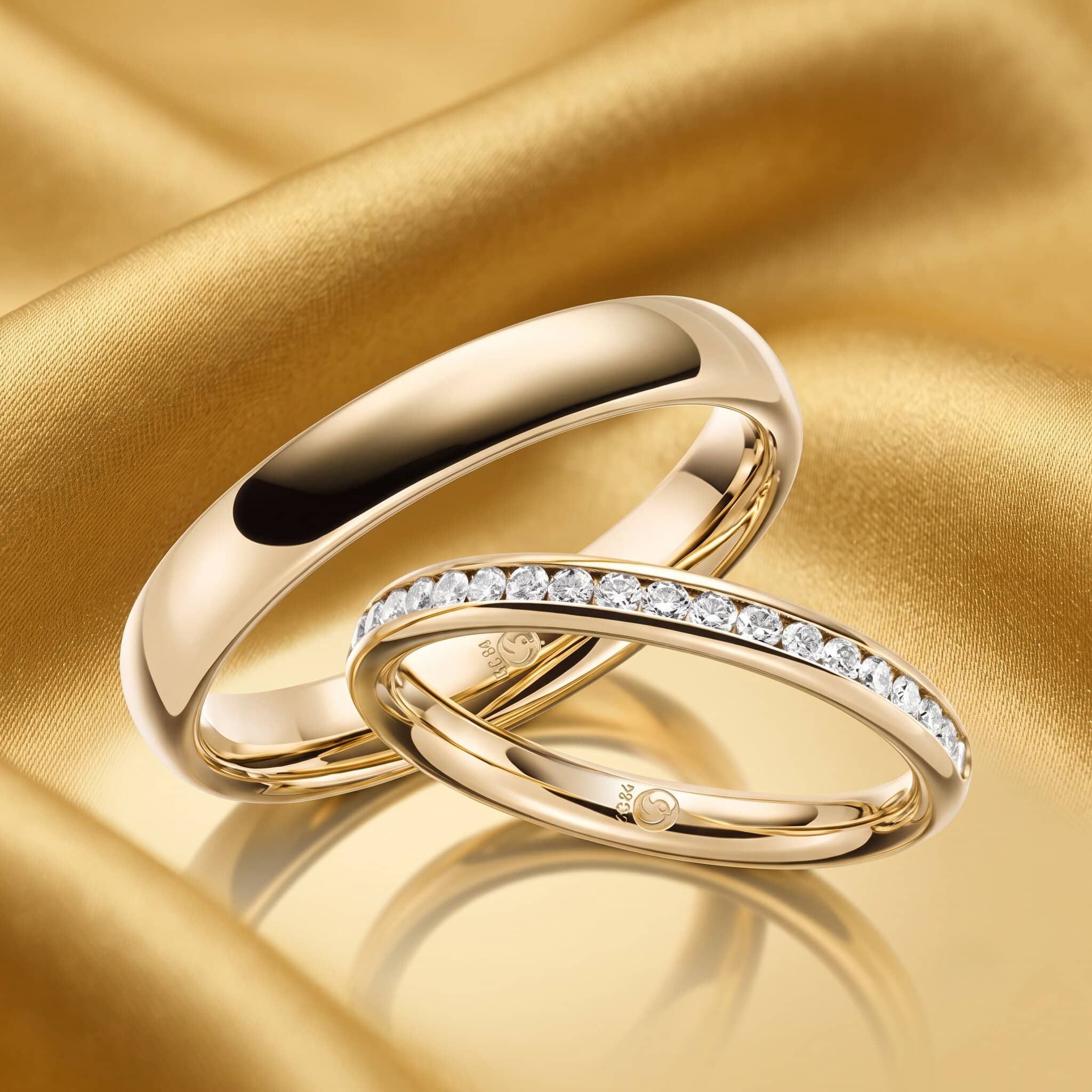 Nahaufnahme zweier luxuriöser Ringe aus Gelbgold, die leicht ineinander verschränkt auf einem fließenden, goldfarbenen Satin- oder Seidenstoff liegen. Der linke Ring ist klassisch schlicht und hochglanzpoliert, der rechte Ring ist mit einer durchgehenden Reihe funkelnder Diamanten im Brillantschliff besetzt. Das Licht erzeugt sanfte Spiegelungen auf dem Gold und dem Stoff.
