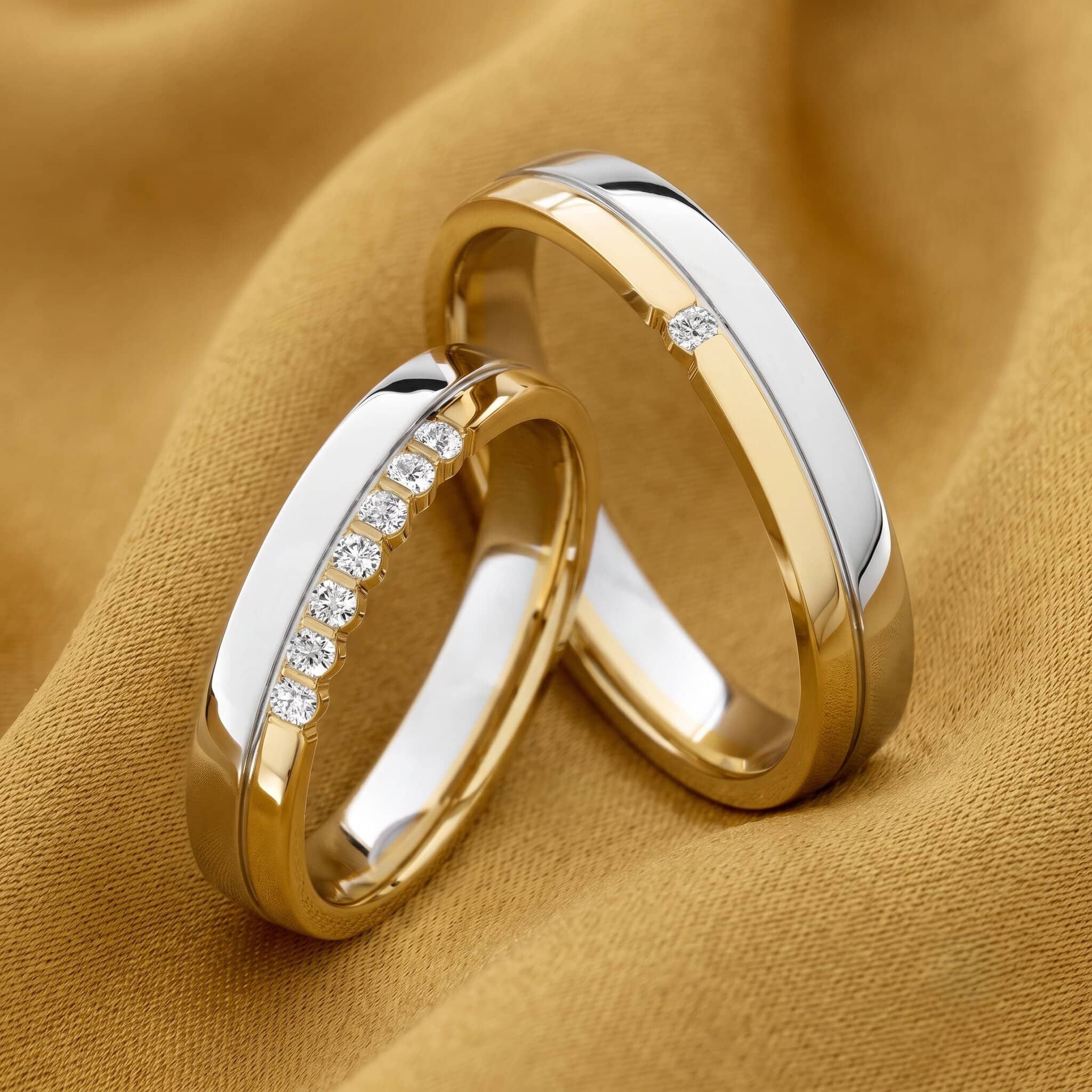 Nahaufnahme zweier ineinander verschlungener Bicolor-Trauringe aus glänzendem Gelbgold und Weißgold, die auf einem weich drapierten, goldfarbenen Stoff liegen. Der linke Ring ist mit einer Reihe kleiner Diamanten in einer eingelassenen Weißgoldschiene verziert. Der rechte Ring kombiniert ebenfalls Gelb- und Weißgold und ist mit einem einzelnen kleinen Diamanten akzentuiert.