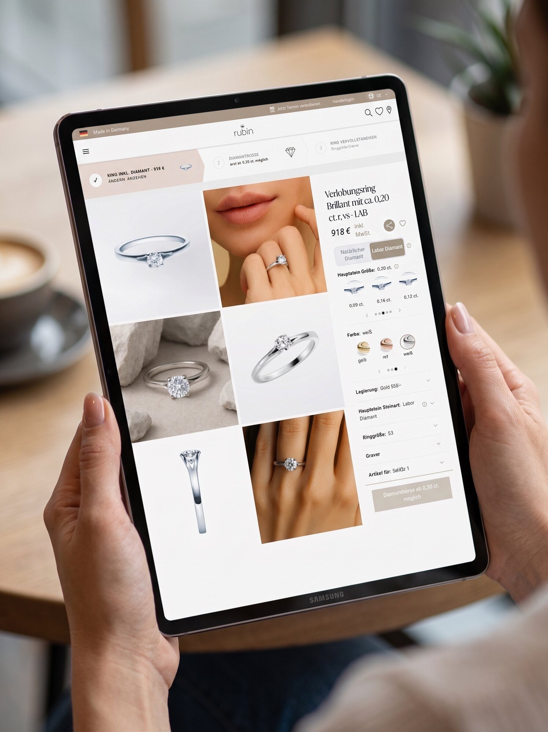 Nahaufnahme einer Person, die ein Samsung-Tablet in den Händen hält. Auf dem Display ist eine Produktseite des Juweliers „rubin“ zu sehen. Die Seite zeigt einen Verlobungsring mit einem 0,20 Karat Lab-Grown-Diamanten für 918 Euro. Es sind verschiedene Ansichten des Rings sowie Auswahloptionen für Karatgewicht, Metallfarbe (Gelb-, Rosé- oder Weißgold) und Legierung abgebildet. Im Hintergrund ist unscharf ein Café-Tisch mit einer Tasse Kaffee zu erkennen.