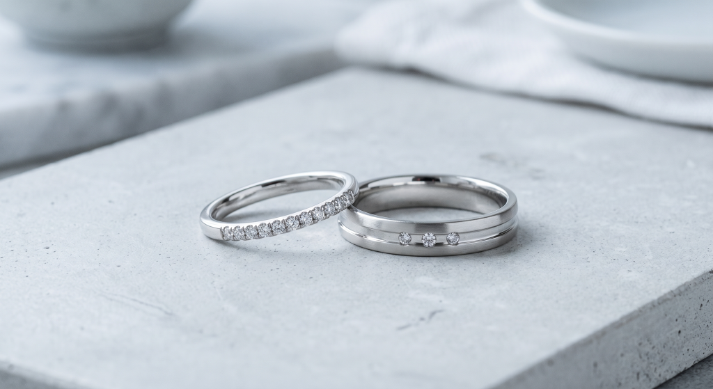 Nahaufnahme von zwei Eheringen aus Weißgold oder Platin auf einer hellgrauen, steinernen Oberfläche. Der linke, zierliche Damenring ist umlaufend mit kleinen Diamanten besetzt. Der rechte, breitere Ring hat eine mattierte Oberfläche, eine feine Rille und drei mittig gefasste Diamanten. Im unscharfen Hintergrund sind helle Keramik- und Stoffsetups zu sehen.